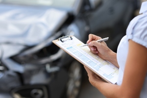 A person writes on a clipboard while inspecting a damaged car with a crumpled front end in the background.