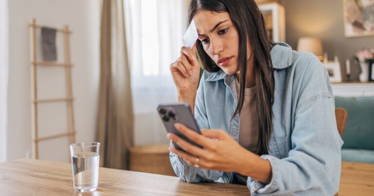 A woman sits at a table holding a phone and a tissue, looking concerned, with a glass of water in front of her in a home setting.