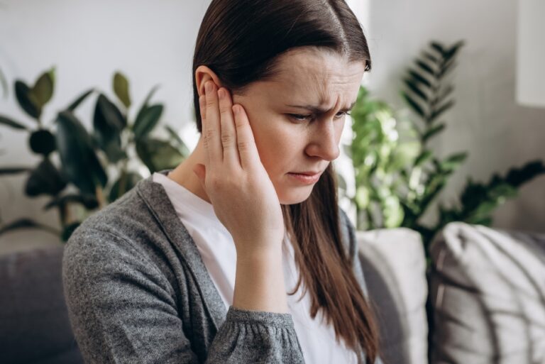 A woman sitting indoors touches her ear and looks uncomfortable, suggesting she may be experiencing ear pain.