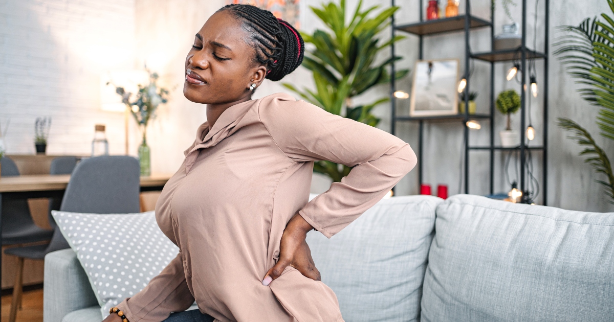 A woman sitting on a couch holds her lower back and closes her eyes, appearing to experience back pain, in a modern living room with plants and shelves in the background.