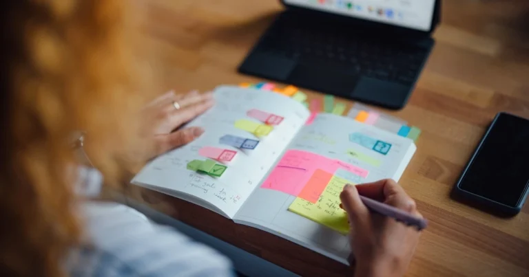 Person writing on a sticky note in a planner with colorful tabs, with a laptop and smartphone on a wooden desk.