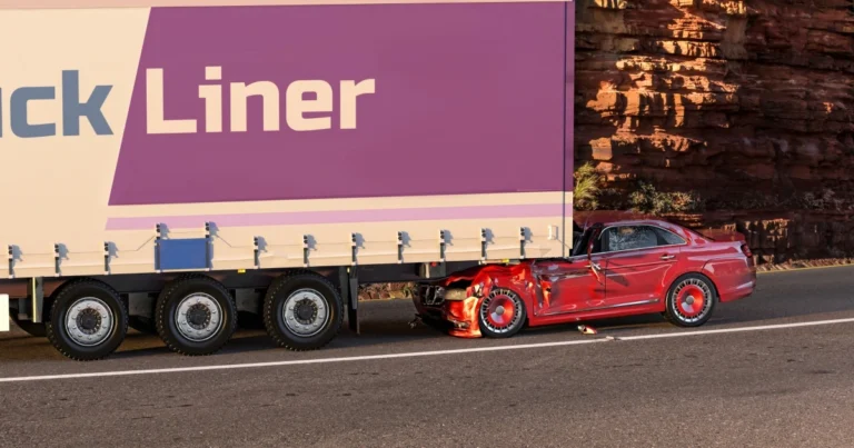 A red car with front-end damage is wedged under the rear of a large truck trailer on the side of a road next to a rocky wall.