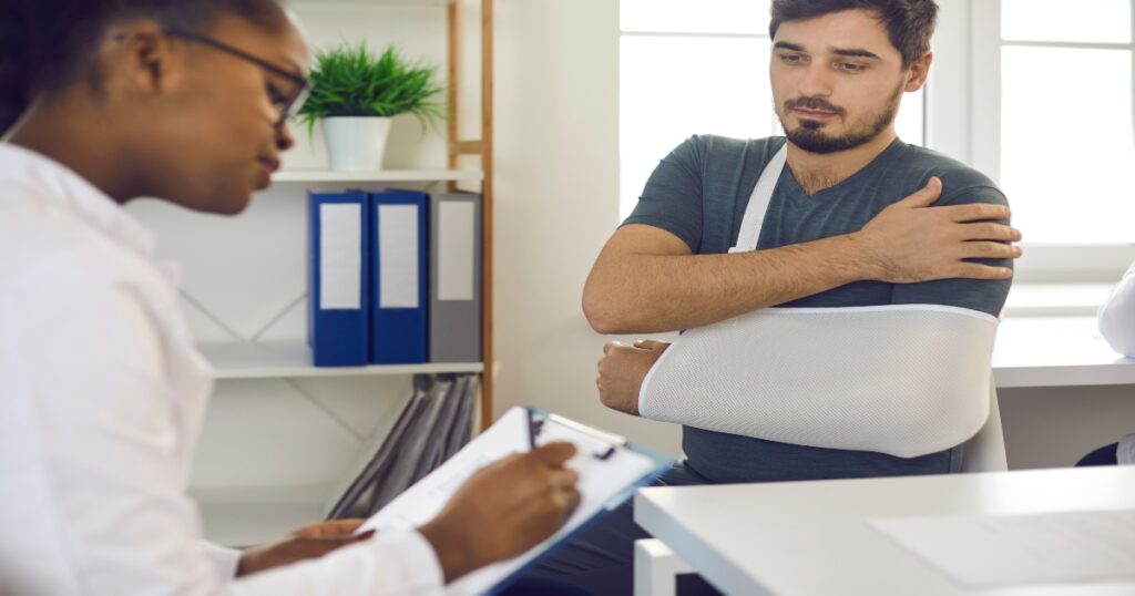 A doctor takes notes while a man with his arm in a sling sits across the desk, holding his injured shoulder.
