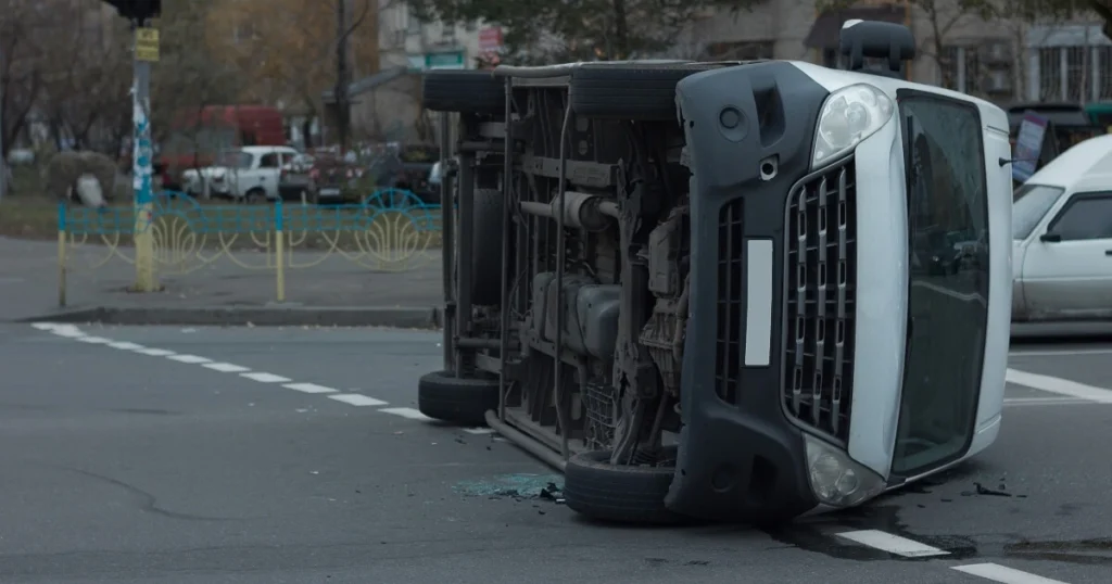 A white van is lying on its side in the middle of an intersection, with broken glass visible on the road.
