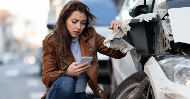 A woman looks worried while inspecting damage to a white car and using her phone, possibly after a car accident on a city street.