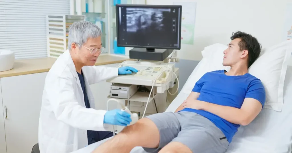 A doctor performs an ultrasound scan on a man's knee as he lies on an examination bed, with the monitor displaying the ultrasound image.
