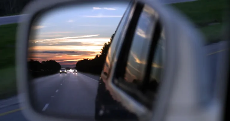 A car’s side mirror reflects a highway at sunset, with headlights of vehicles visible behind and the sky displaying orange and purple hues.