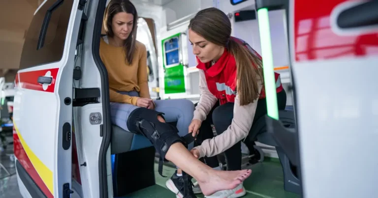 A paramedic fits a knee brace on a woman’s leg inside an ambulance. The woman sits with her injured leg extended while the paramedic carefully adjusts the brace.