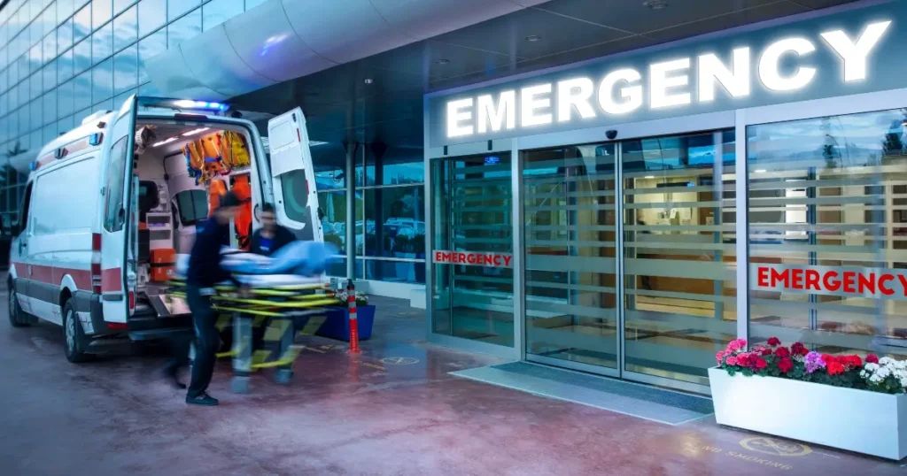 Paramedics wheel a patient on a stretcher from an ambulance into a hospital emergency room entrance at night.
