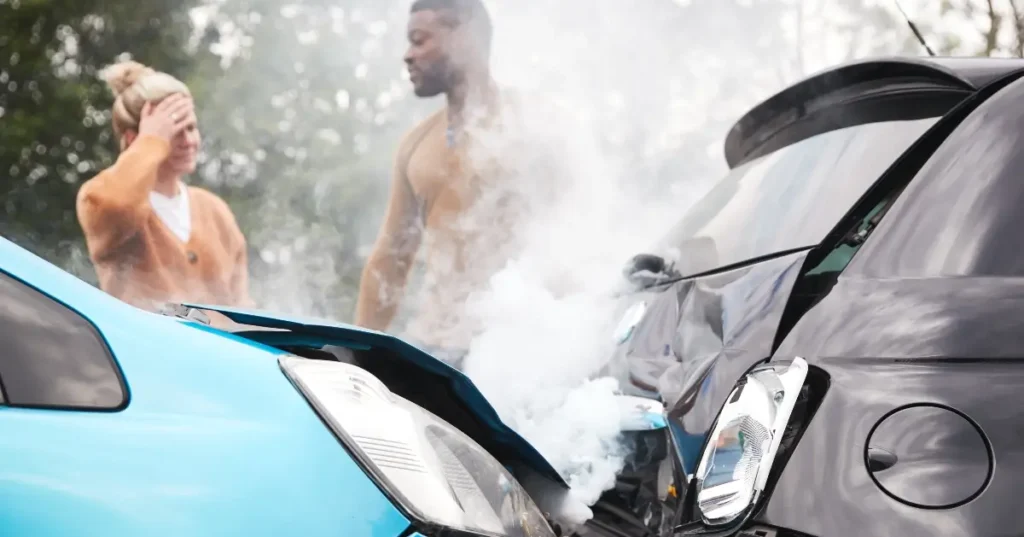 Two damaged cars after a collision, with smoke rising from the impact. Two people stand nearby, one holding their head, both appearing concerned.