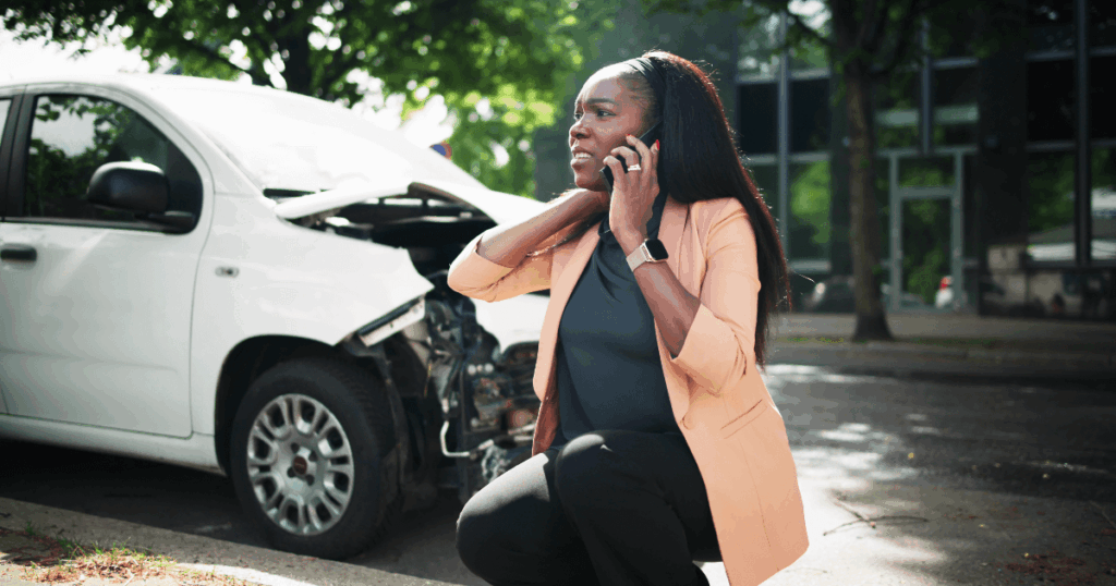 A woman kneels on the street while making a phone call, with a damaged white car and trees in the background.