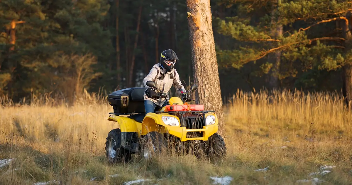 Person wearing a helmet and jacket rides a yellow all-terrain vehicle (ATV) through a grassy, wooded area with trees in the background.