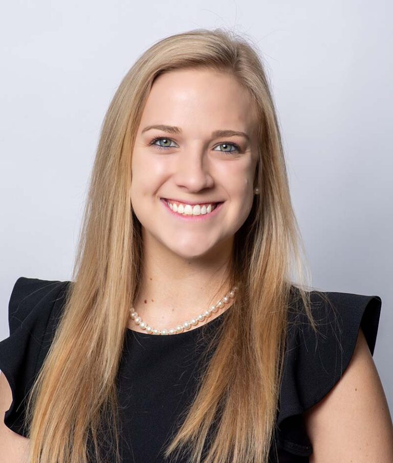 A woman with long blonde hair, wearing a black top and a pearl necklace, smiles at the camera against a plain light background.