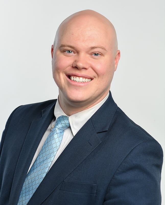 A bald man in a dark suit, light blue dress shirt, and patterned light blue tie smiles at the camera against a plain, light background.