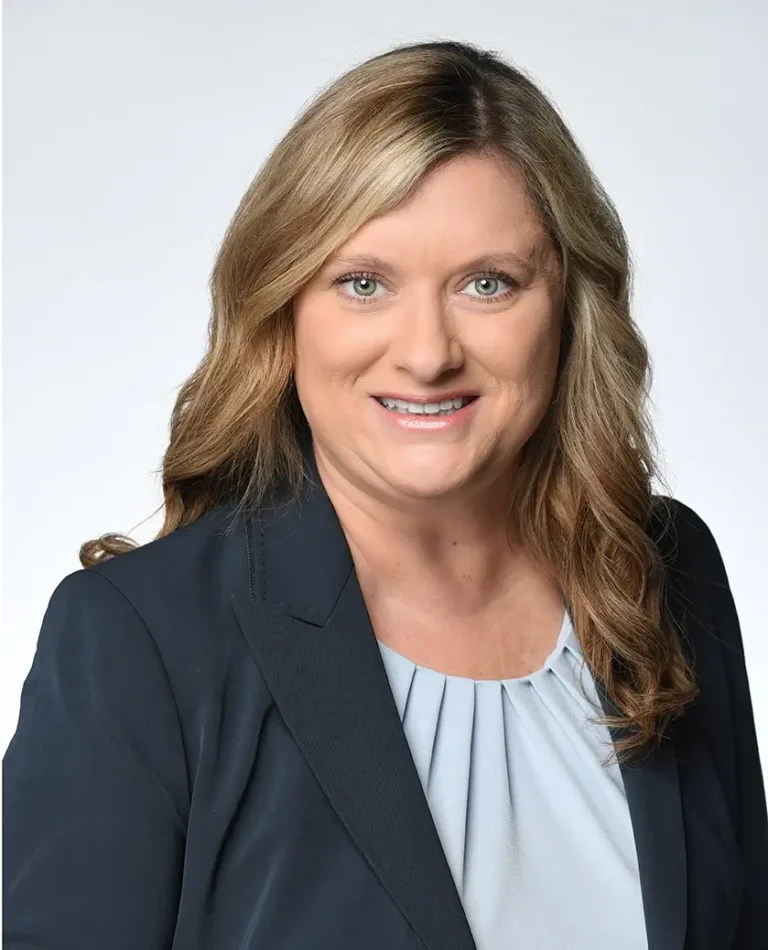 A woman with wavy blond hair wearing a navy blazer and light blouse smiles at the camera against a plain light background.