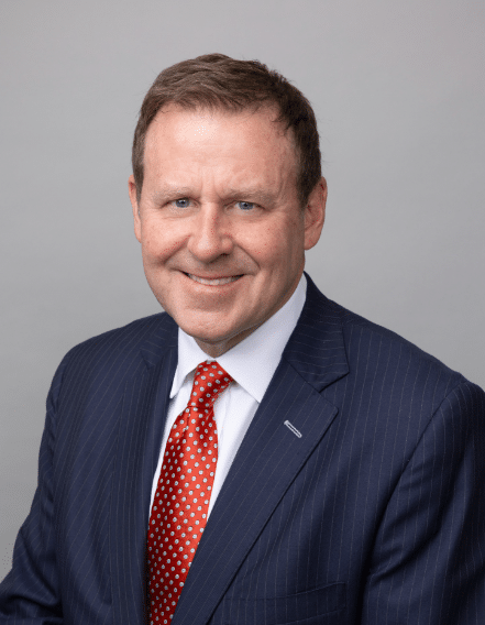 A man in a navy pinstripe suit, white dress shirt, and red polka dot tie poses for a professional headshot against a plain gray background.