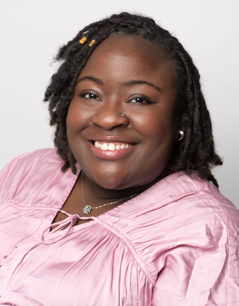 A woman with medium-dark skin and short, beaded dreadlocks smiles at the camera. She is wearing a pink blouse and layered necklaces.