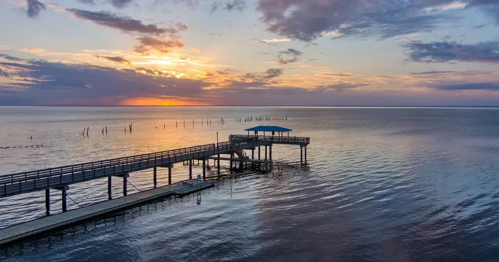 A long wooden pier with a covered section extends over calm water at sunset, with clouds partially covering the sky.
