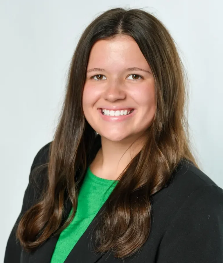 A woman with long brown hair wearing a black blazer over a green top, smiling at the camera against a plain light background.