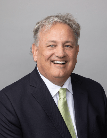 Smiling man with gray hair wearing a dark suit, white shirt, and light green tie, posed in front of a plain gray background.