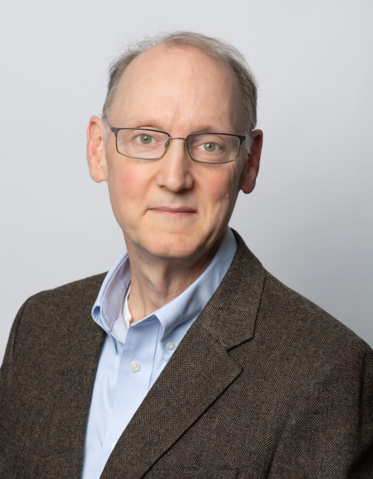 A middle-aged man with glasses, short graying hair, and a brown blazer over a light blue shirt poses in front of a plain light background.