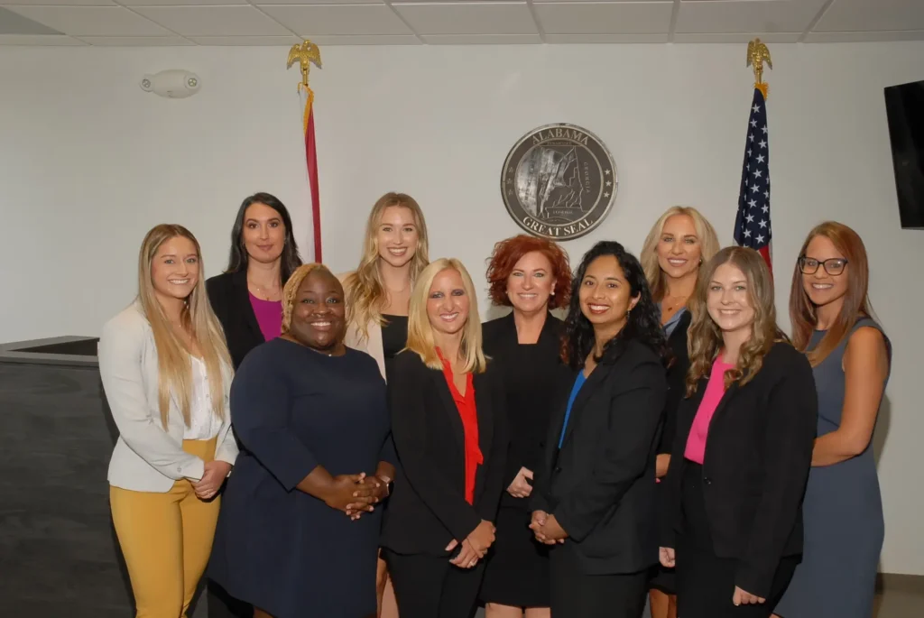 A group of ten women in business attire pose for a photo in front of the Alabama Great Seal and two flags inside a formal room.