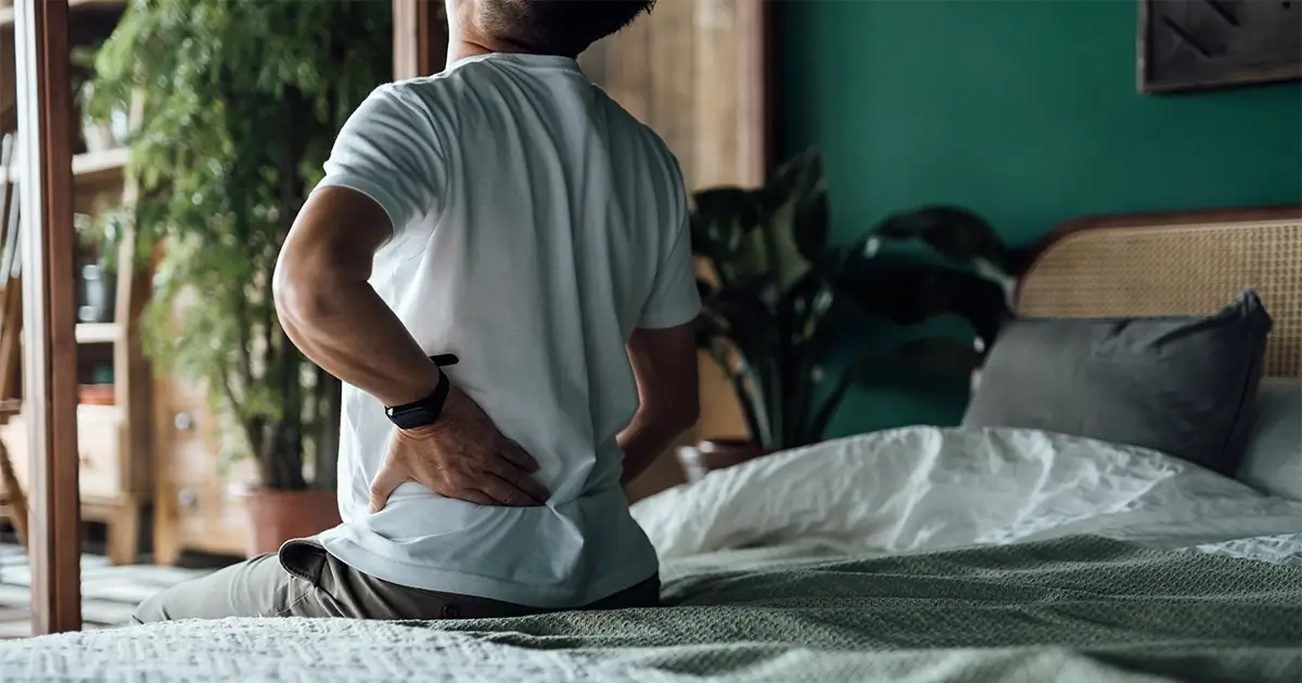 A person sits on the edge of a bed, holding their lower back with one hand, appearing to experience back pain.