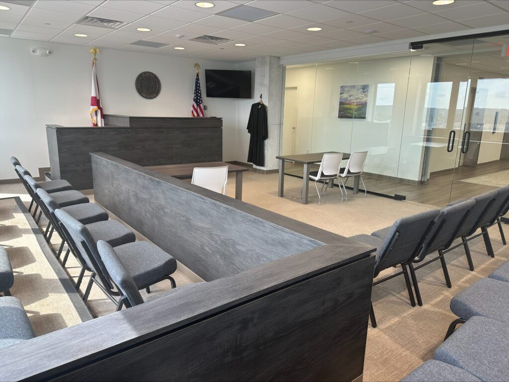 Modern courtroom with empty chairs, a judge’s bench, flags, and a table with two chairs next to a glass wall; a black robe hangs on the wall.
