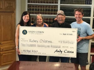 Four people stand indoors holding a large ceremonial check made out to Rickey Childress from Andy Citrin Injury Attorneys.