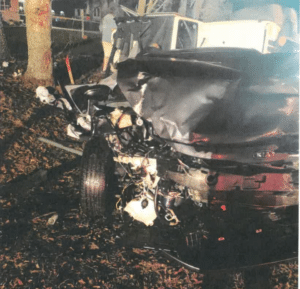 A heavily damaged vehicle is shown after a severe collision with a tree at night; debris and wreckage are scattered around the crash site.
