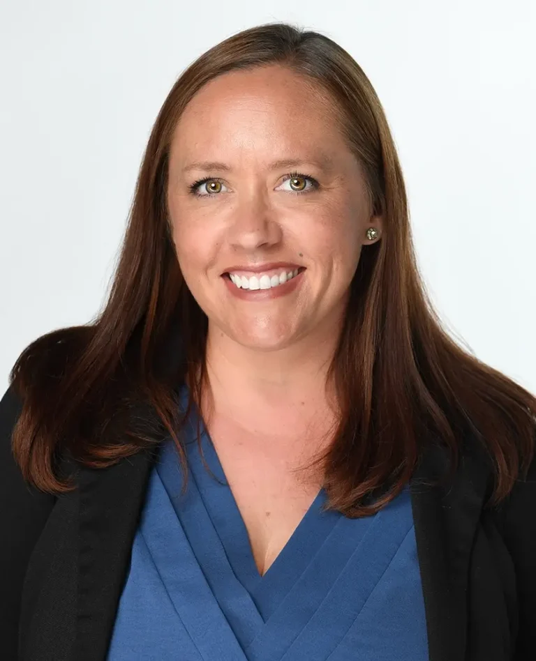 Woman with long brown hair, wearing a blue blouse and black blazer, smiles at the camera against a plain white background.