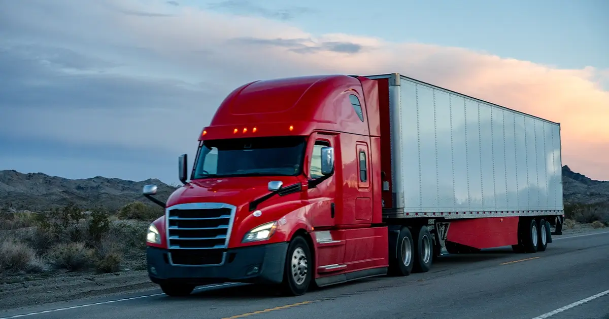 A red semi-truck with a white trailer drives on a highway through a desert landscape at sunset.