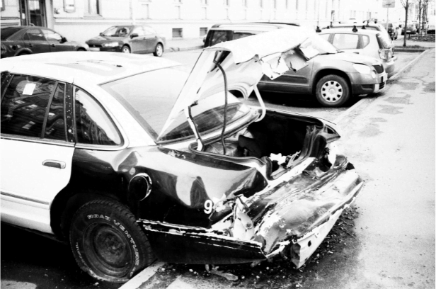 A car with severe damage to its rear end is parked on a street beside other vehicles. The trunk is crumpled and open, and the license plate is missing.