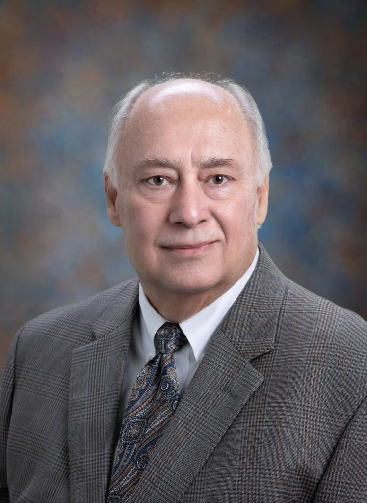 An older man with short white hair wearing a plaid suit jacket, white shirt, and patterned tie poses for a formal portrait against a studio background.