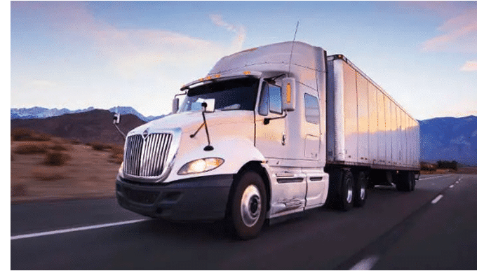 A silver semi-truck with a trailer drives on a highway with mountains and a sunset in the background.