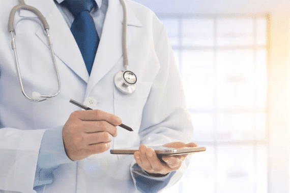 A doctor in a white coat with a stethoscope uses a stylus on a digital tablet in a bright, modern setting.