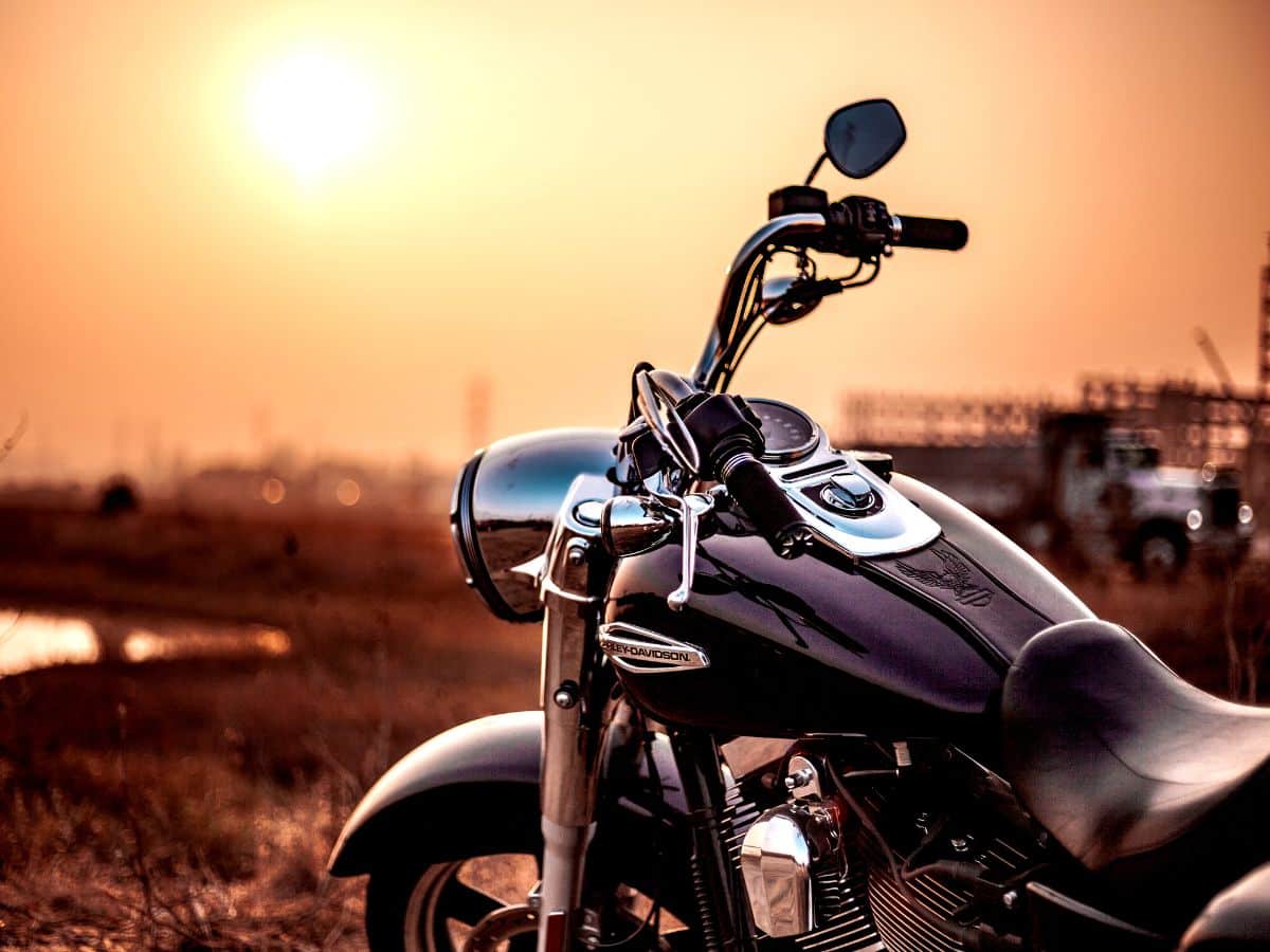 Close-up of a black motorcycle parked outdoors at sunset, with sunlight reflecting off its chrome parts and a blurred field and truck in the background.