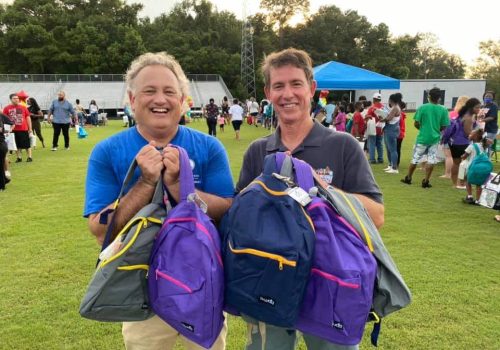 Two men standing on grass hold colorful backpacks at an outdoor event, with a crowd of people and tents visible in the background.
