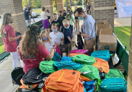A group of adults and children, some wearing masks, gather around a table with colorful backpacks outside a building.