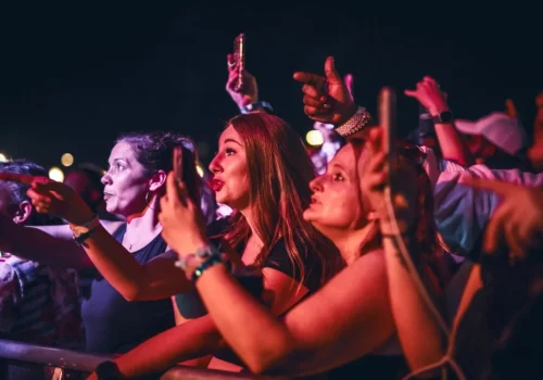 A group of people at a concert stand near the stage barrier, some holding phones and pointing, illuminated by colorful stage lights at night.