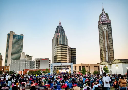 A large crowd gathers for an outdoor event in front of tall buildings in a city downtown area during daylight.