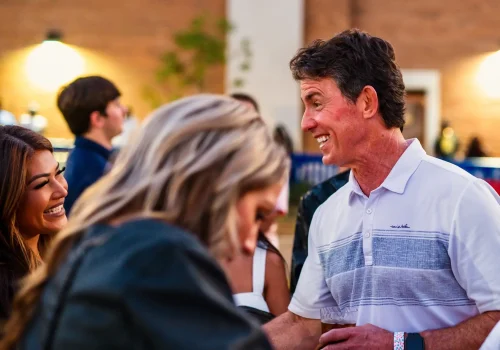 A man in a white polo shirt smiles and talks with two women at an outdoor gathering; blurred people and a brick wall are in the background.