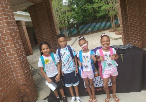 Four children with backpacks stand outside near a brick building, smiling at the camera. One child is wearing a face mask. A table with a "backpack" sign is behind them.