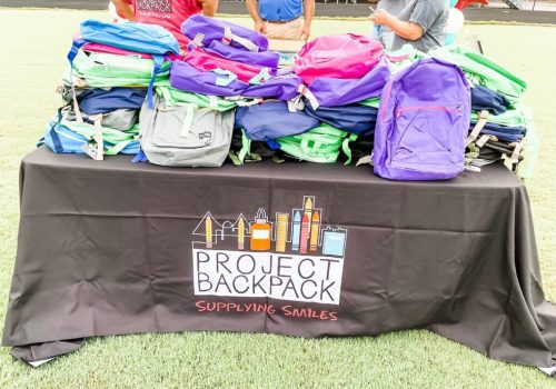 A table with a "Project Backpack" tablecloth holds stacks of colorful backpacks; several people stand behind the table on a grassy field.