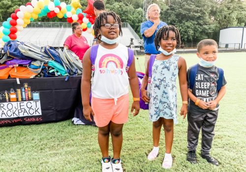 Three children with backpacks stand on grass in front of a table labeled "Project Backpack" with school supplies, balloons, and adults in the background.