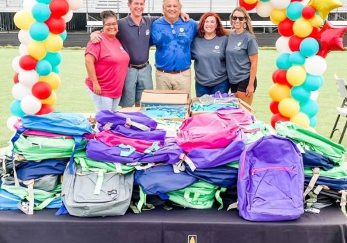 Five people stand behind a table with stacked backpacks and a "Project Backpack" banner, under a colorful balloon arch on a field with empty bleachers in the background.