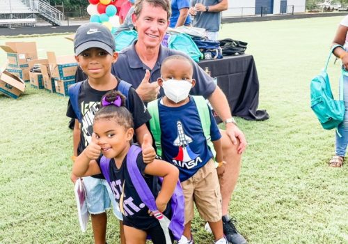 A man kneels on grass with three young children wearing backpacks at an outdoor event; balloons and tables are visible in the background.