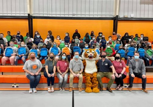 A group of students with blue backpacks and masks sit in bleachers behind staff members and a person in a tiger mascot costume, inside a gymnasium with orange and black walls.