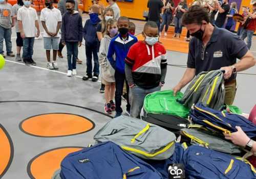 Students stand in line in a gymnasium as adults distribute backpacks from a large pile on a table. Some people are wearing masks.